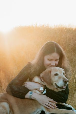 Young woman smiling and hugging her golden Labrador dog in a sunlit field of golden grass at sunset.