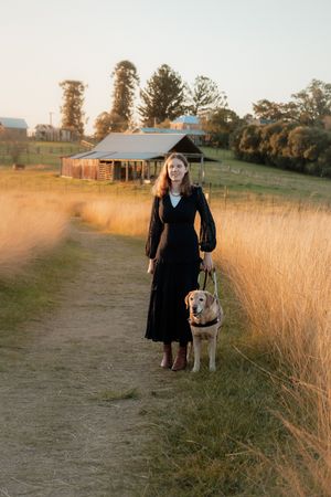 Woman in black dress walking golden Labrador on rural path at sunset with rustic barn and golden grass fields.