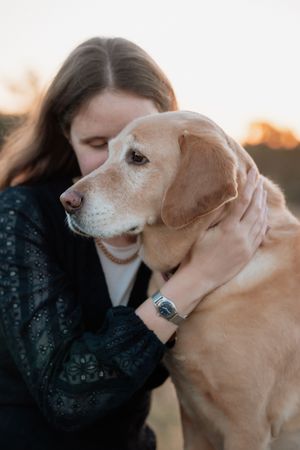 A young woman in a black lace jacket tenderly embraces her golden Labrador dog outdoors at sunset.