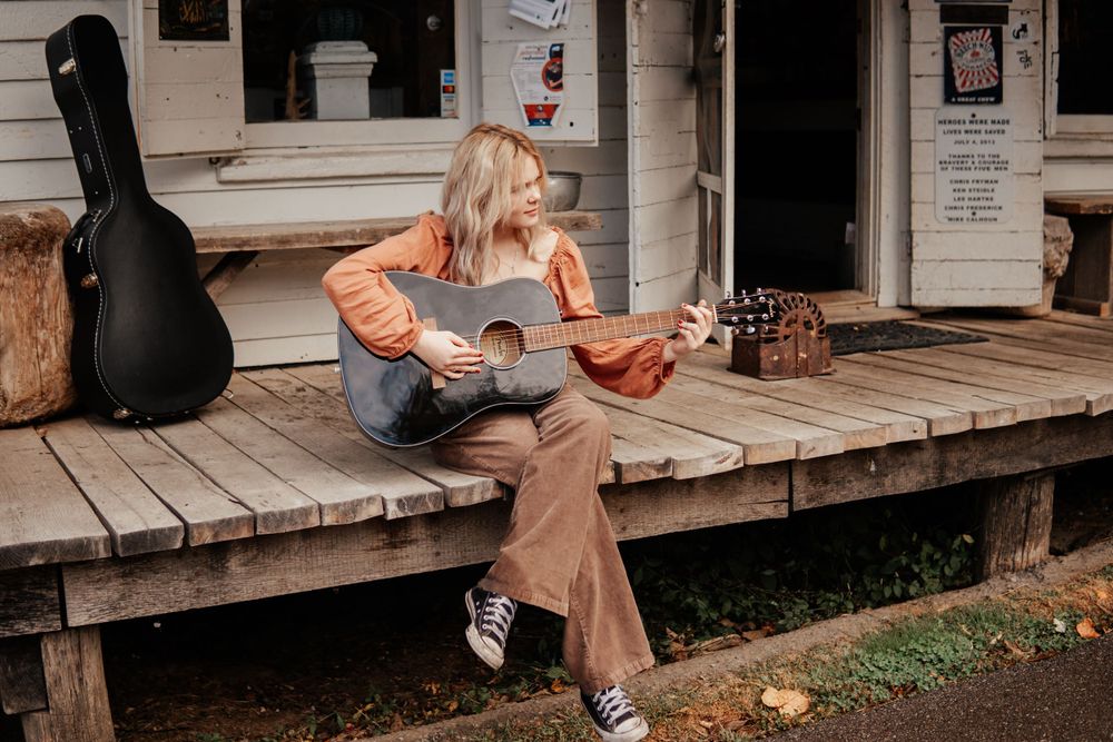 A musician plays acoustic guitar while sitting on a wooden porch with a guitar case nearby.