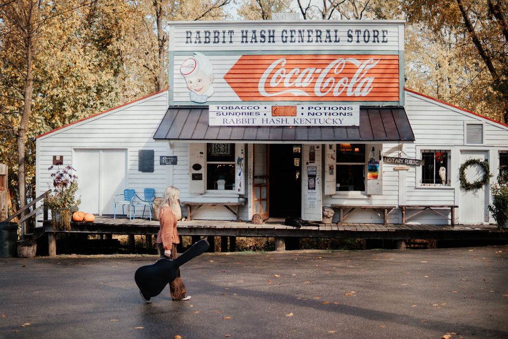 A person with luggage stands in front of a vintage general store with Coca-Cola signage.