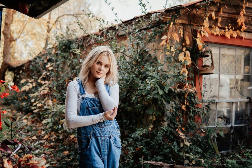 A woman in overalls and white long sleeve shirt stands against a rustic backdrop with green foliage.