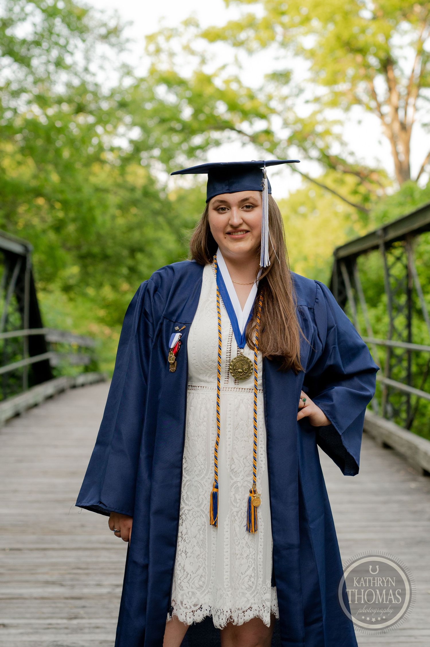 Graduate in cap and gown posing on wooden bridge with trees in background.