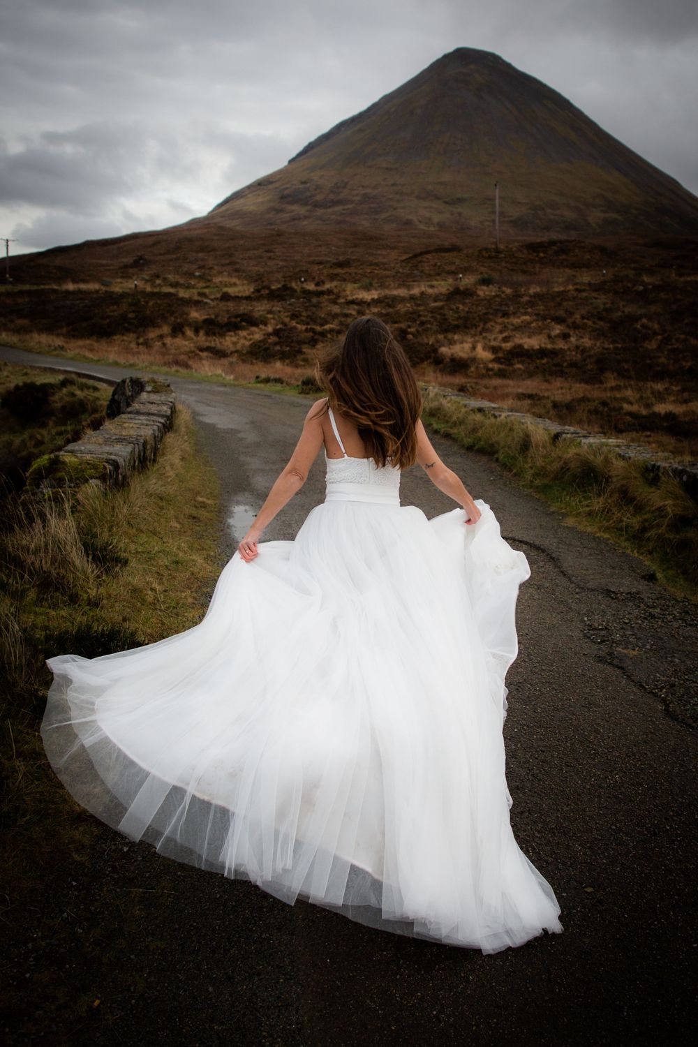 A person in a flowing white wedding dress twirls on a remote road with a dramatic mountain landscape in the background.