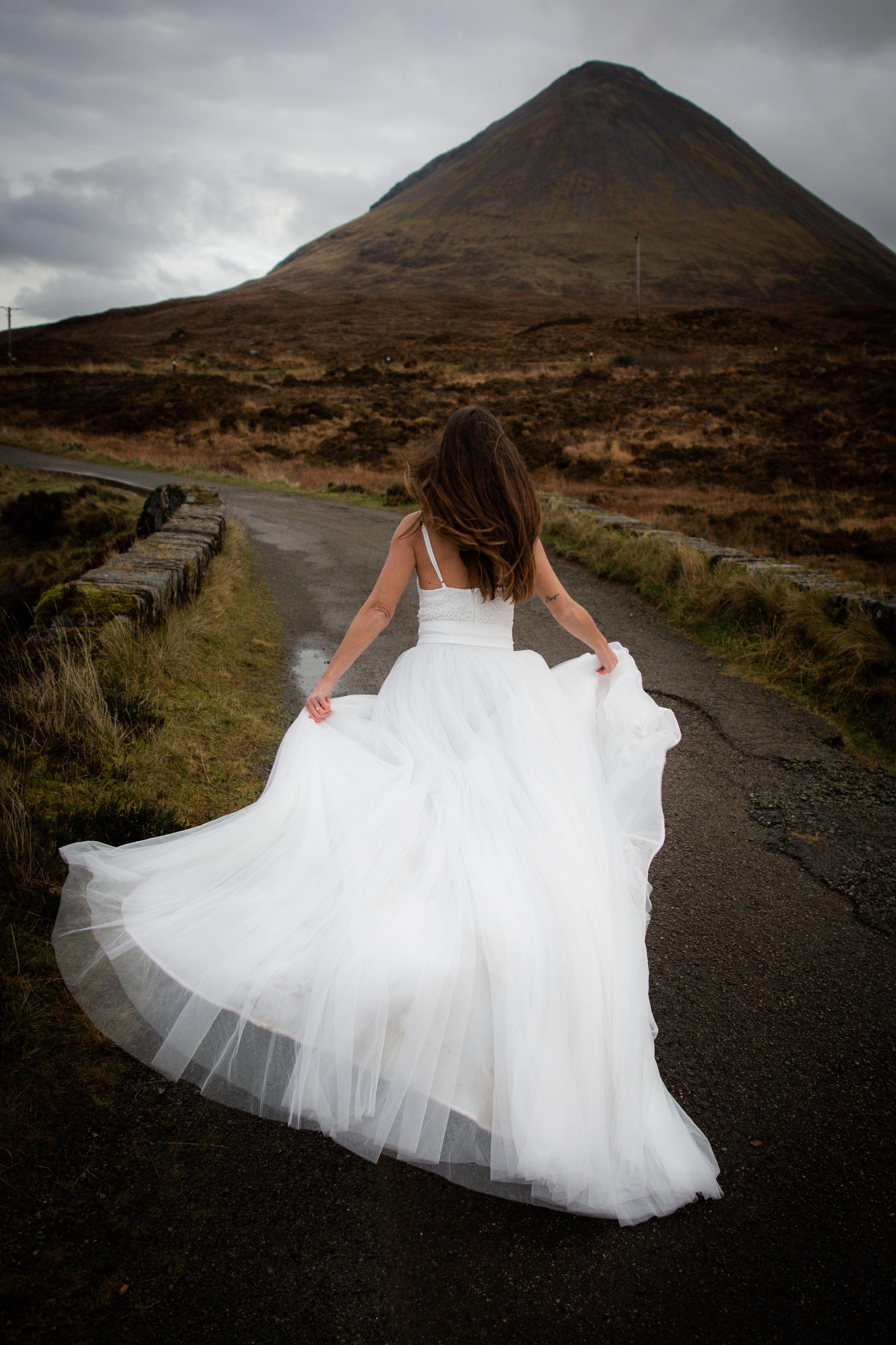 A person in a flowing white wedding dress twirls on a remote road with a dramatic mountain landscape in the background.