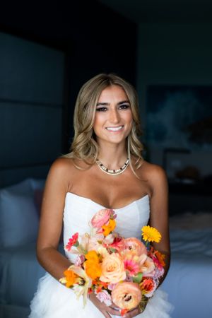 A bride in a white strapless gown holds a vibrant bouquet of coral, orange and pink flowers while smiling.