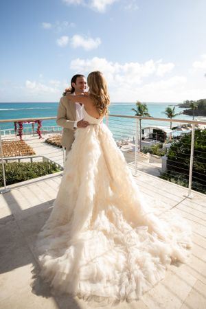 Newlywed couple embrace on a tropical beachfront balcony with turquoise ocean views and palm trees in the background.