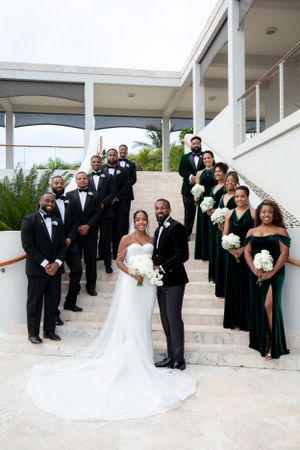 Elegant wedding party poses on white stairs with bridesmaids in dark green dresses and groomsmen in black tuxedos.