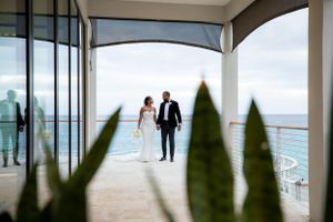 A couple in formal attire stands framed by a curved balcony with ocean views, seen through decorative shrubs in foreground.
