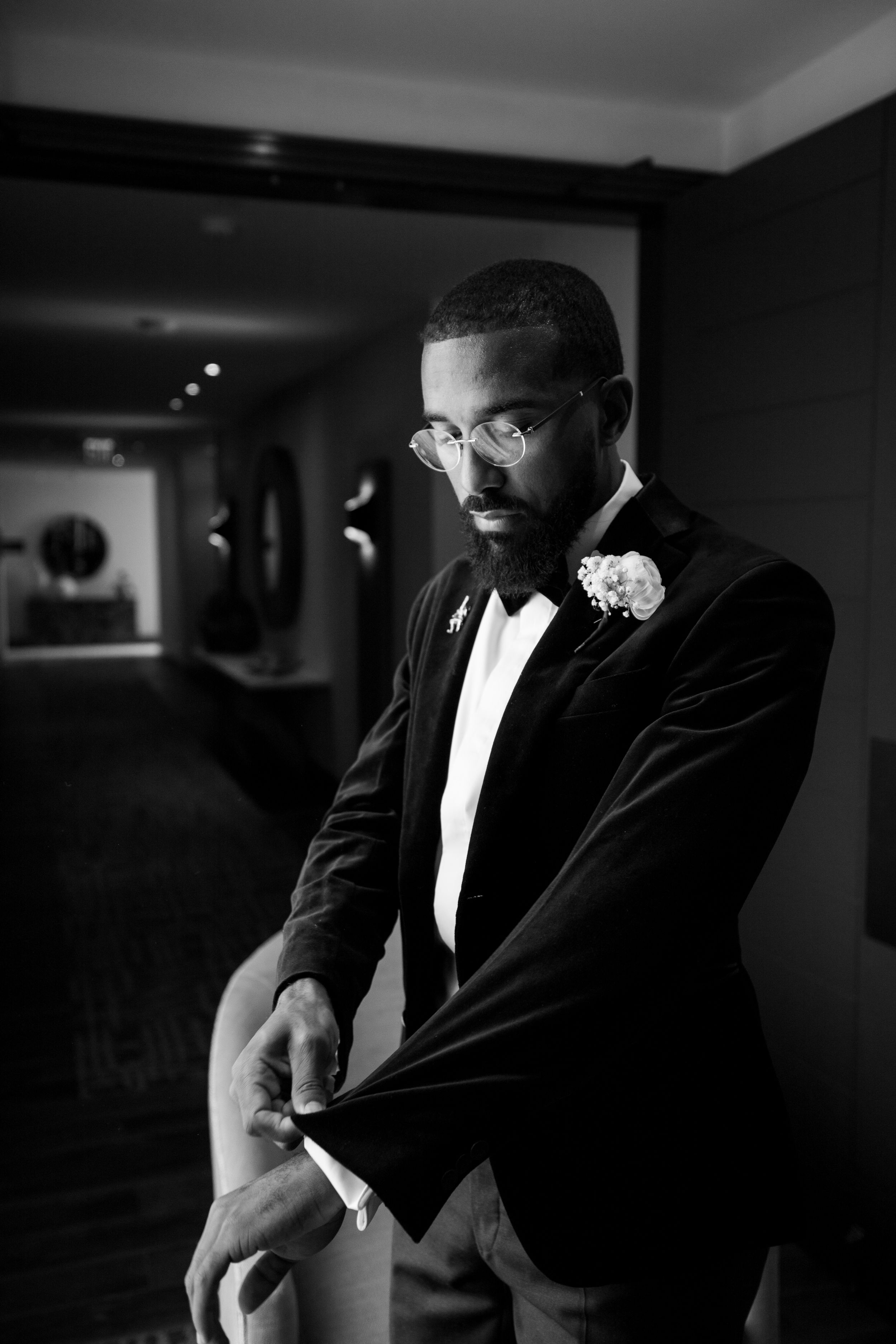 A black and white formal portrait of a groom in a velvet tuxedo with glasses and boutonniere in a dark hallway.