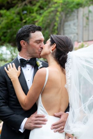 A tender wedding kiss between newlyweds, with the bride in a backless white dress and groom in classic tuxedo.