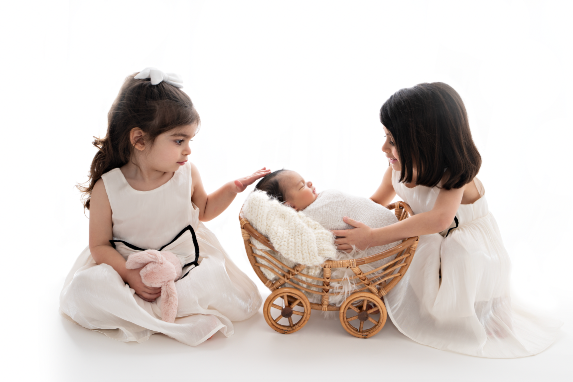 Two young girls in white dresses admire a sleeping newborn baby in a wicker pram during a studio photo session.
