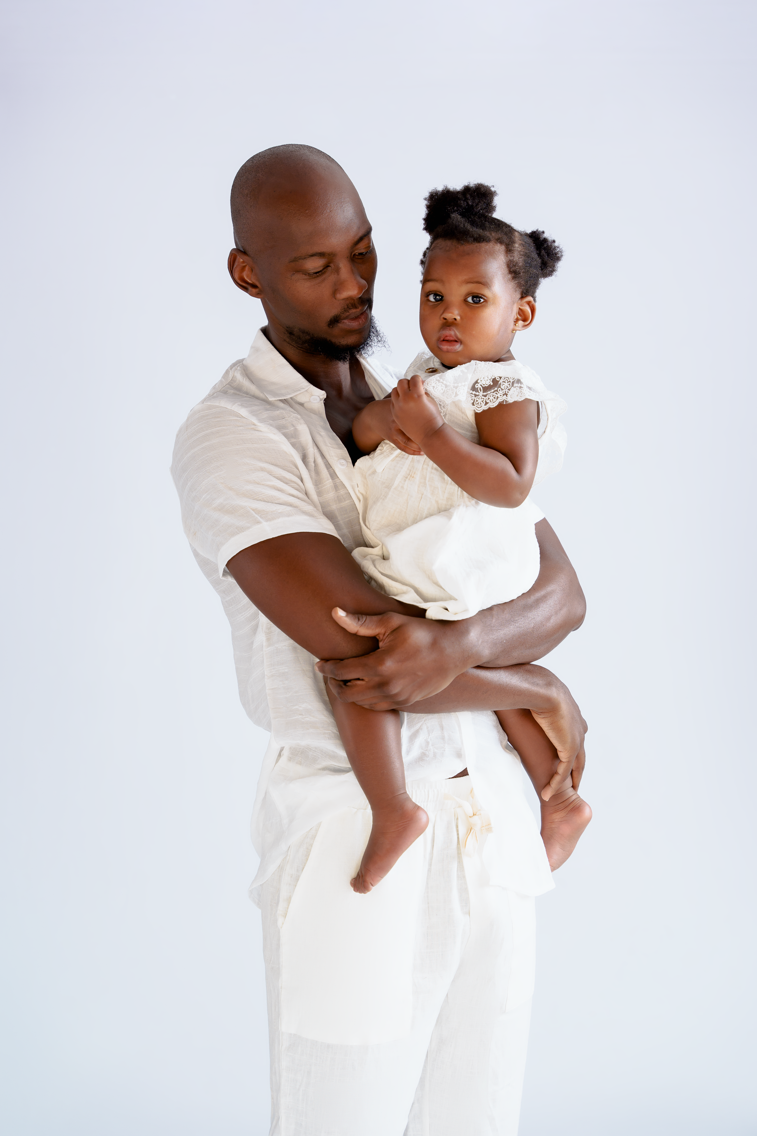 Parent in white outfit holding small child wearing white dress against light background.