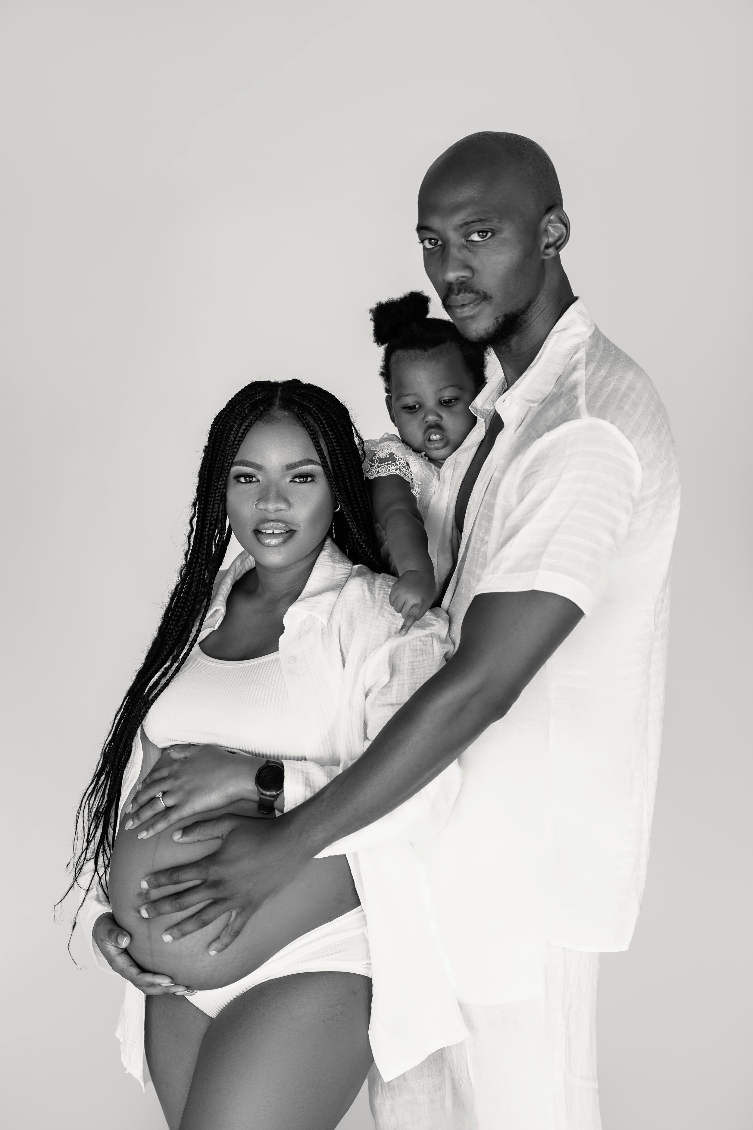 Intimate black and white maternity photo of a couple in white clothing embracing with a small child.