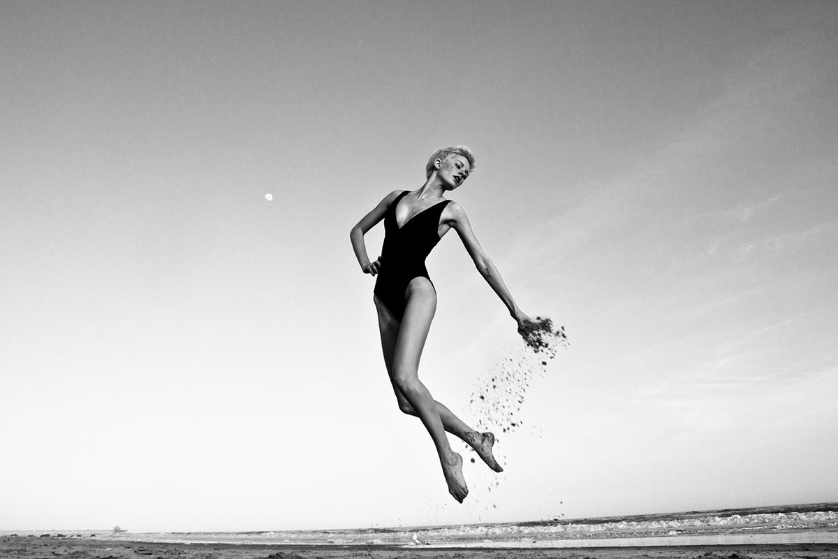 Graceful dancer in black leotard leaps through the air on a beach in dramatic black and white photography.
