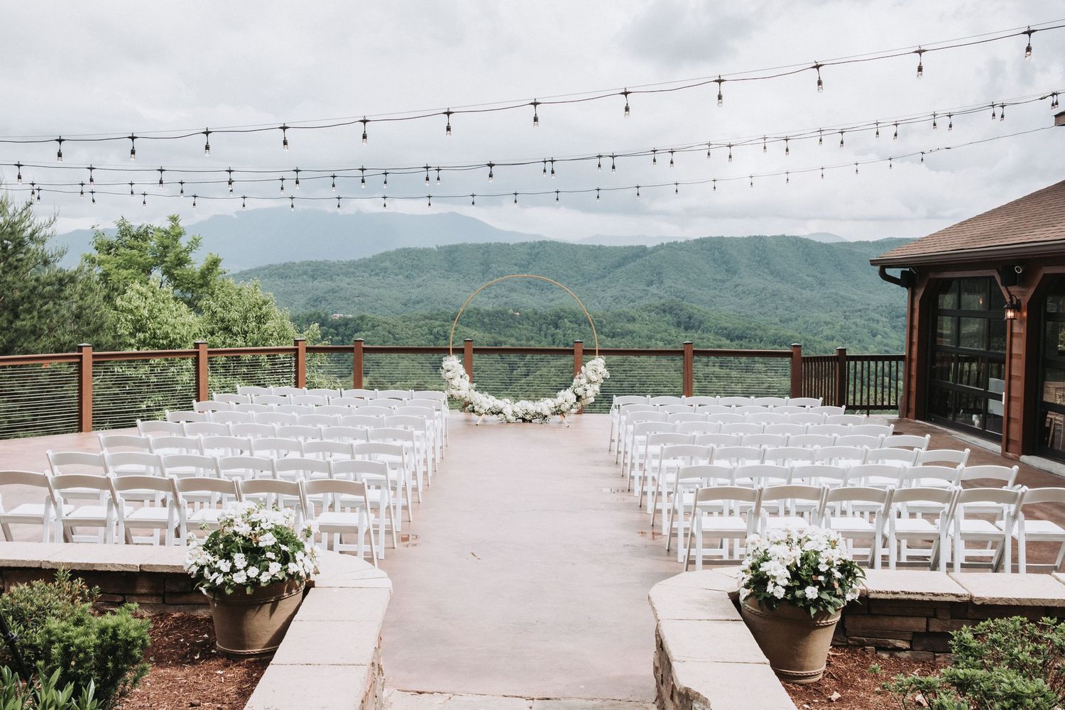 A mountain view wedding ceremony setup with white chairs and string lights on an outdoor deck overlooking rolling hills.