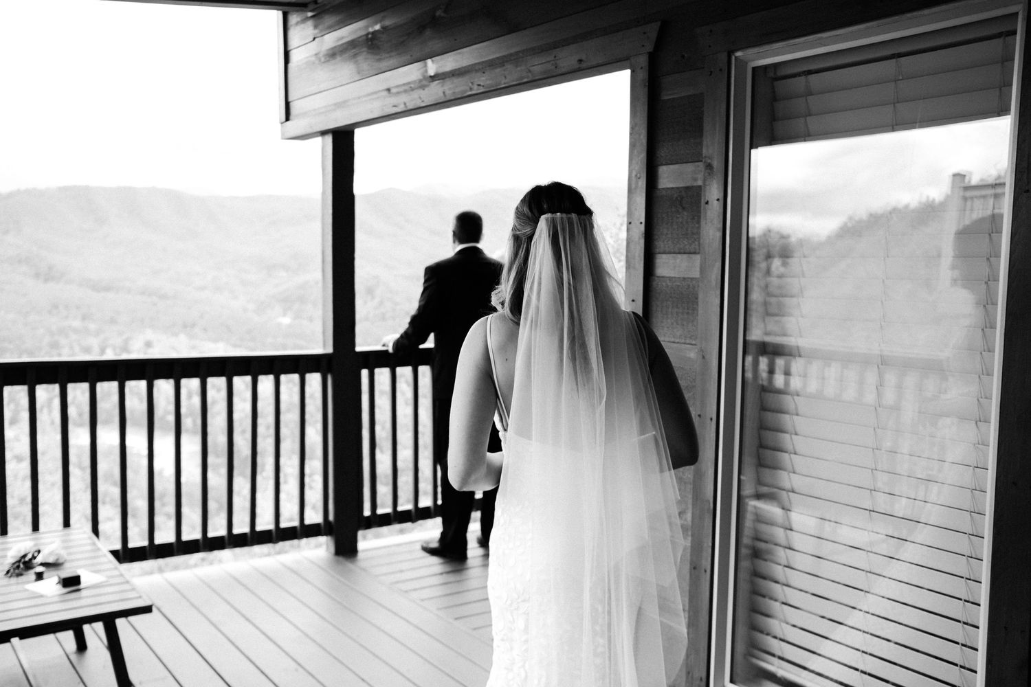 A black and white wedding photo captures a bride in flowing veil approaching her partner on a covered deck overlooking hills.