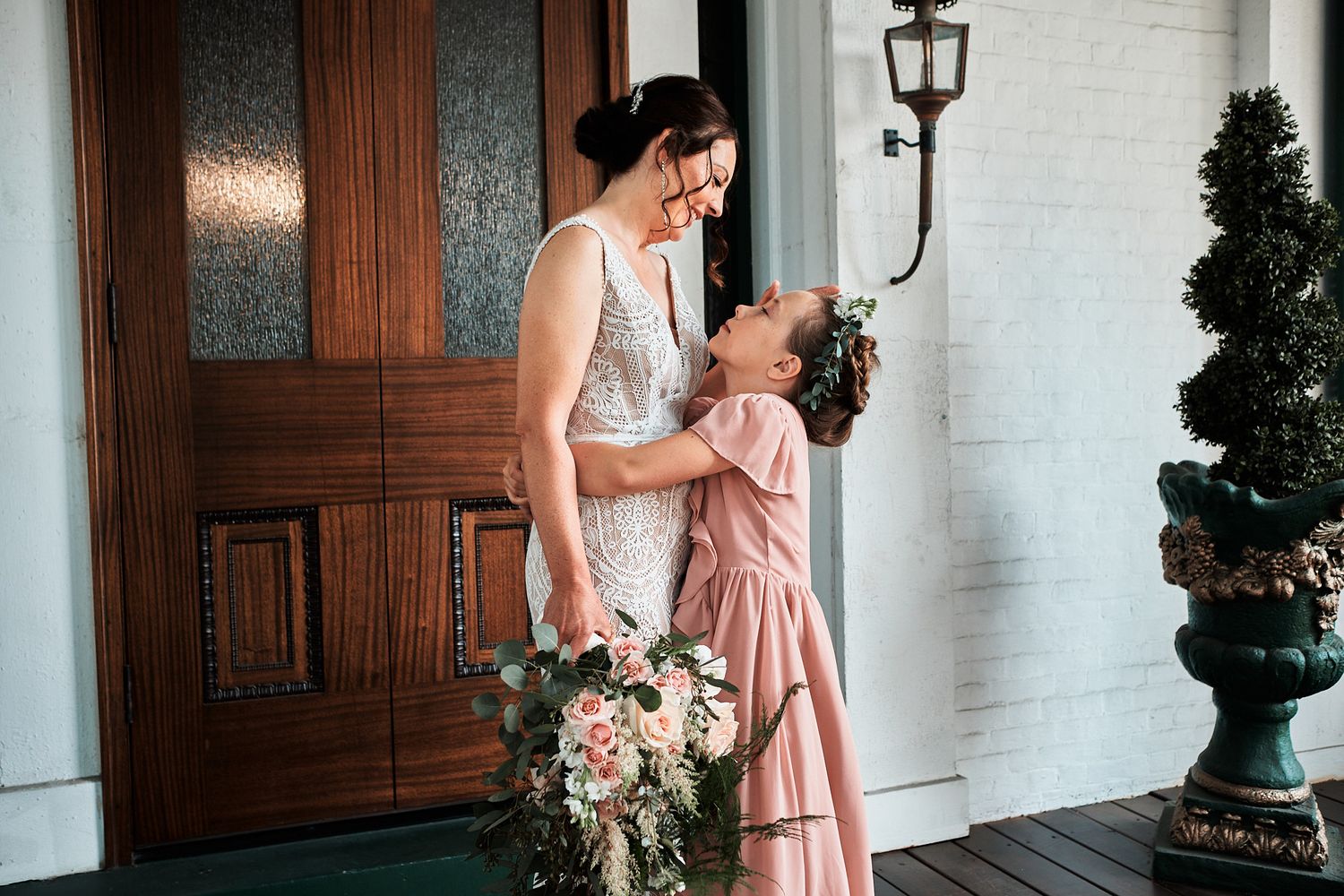 A tender moment between bride and flower girl on a white porch with floral bouquet.