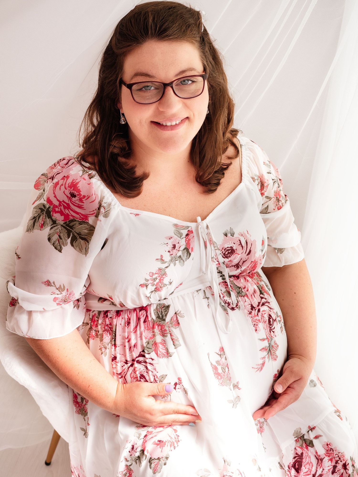 A beautiful pregnant woman in white dress with pink roses sits and smiles at the photographer