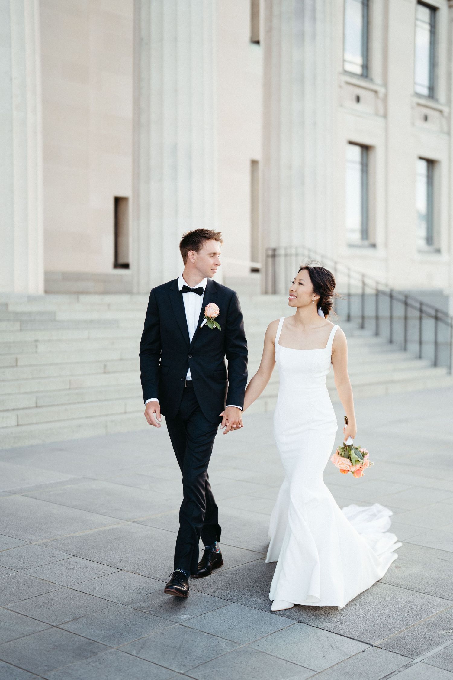 A stylish couple walks hand in hand along grand classical columns in formal wedding attire.
