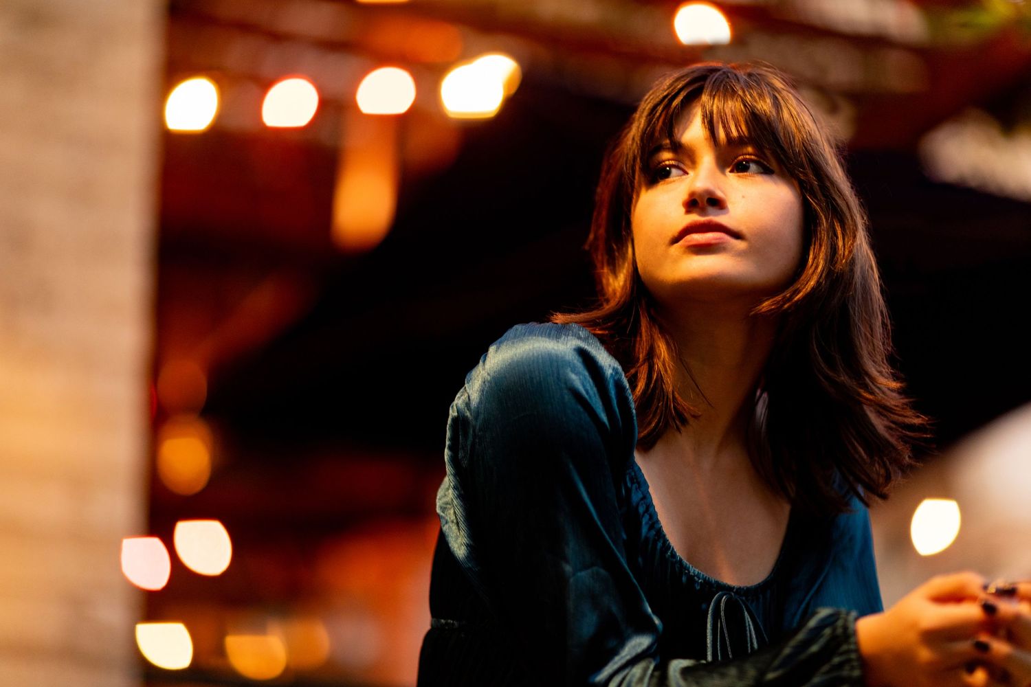 A high school senior in dark clothing looking upward with warm bokeh lights in the background creating moody ambiance.