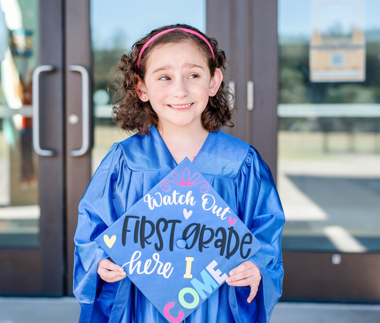 Graduate in blue gown points to decorated cap with colorful lettering outside school building.