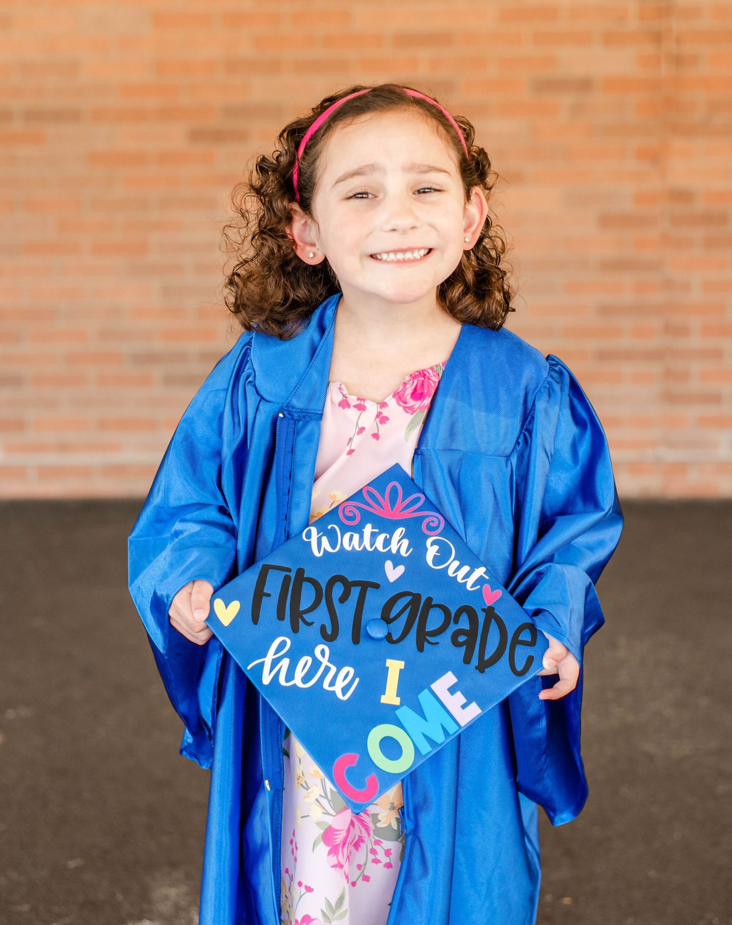 Graduate wearing floral dress under blue graduation gown poses in front of brick wall.
