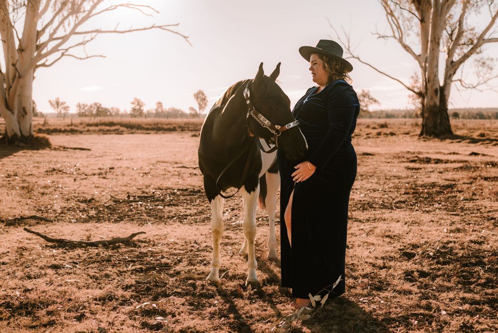 A horse and figure in dark clothing stand together in a rural Australian landscape at sunset.