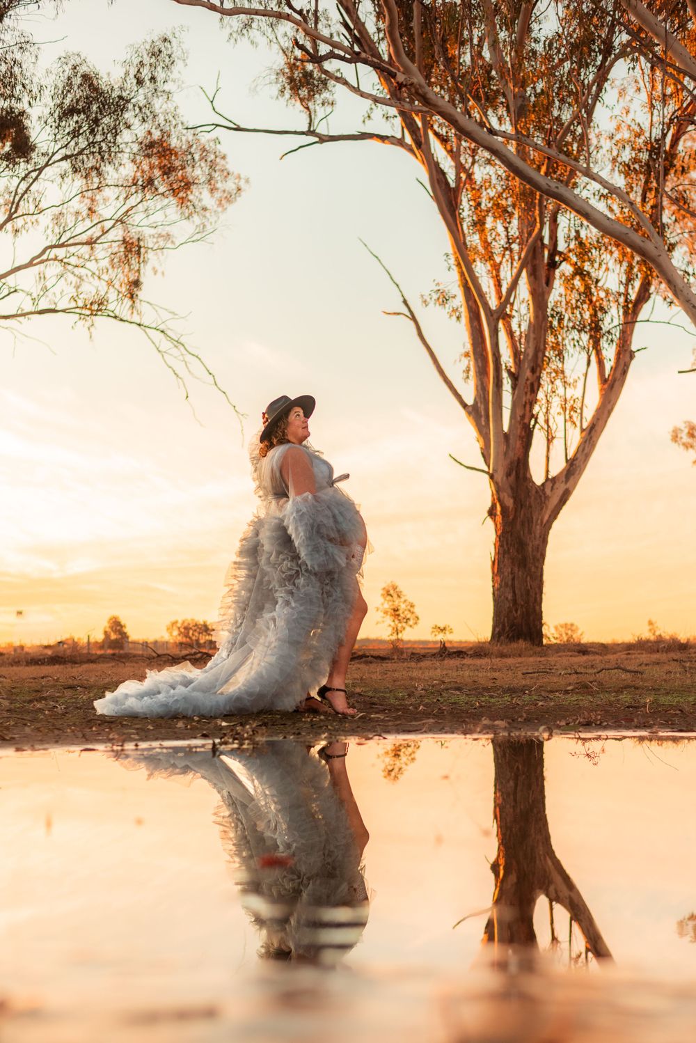 A figure in a flowing gown stands reflected in still water at sunset with a gum tree backdrop.