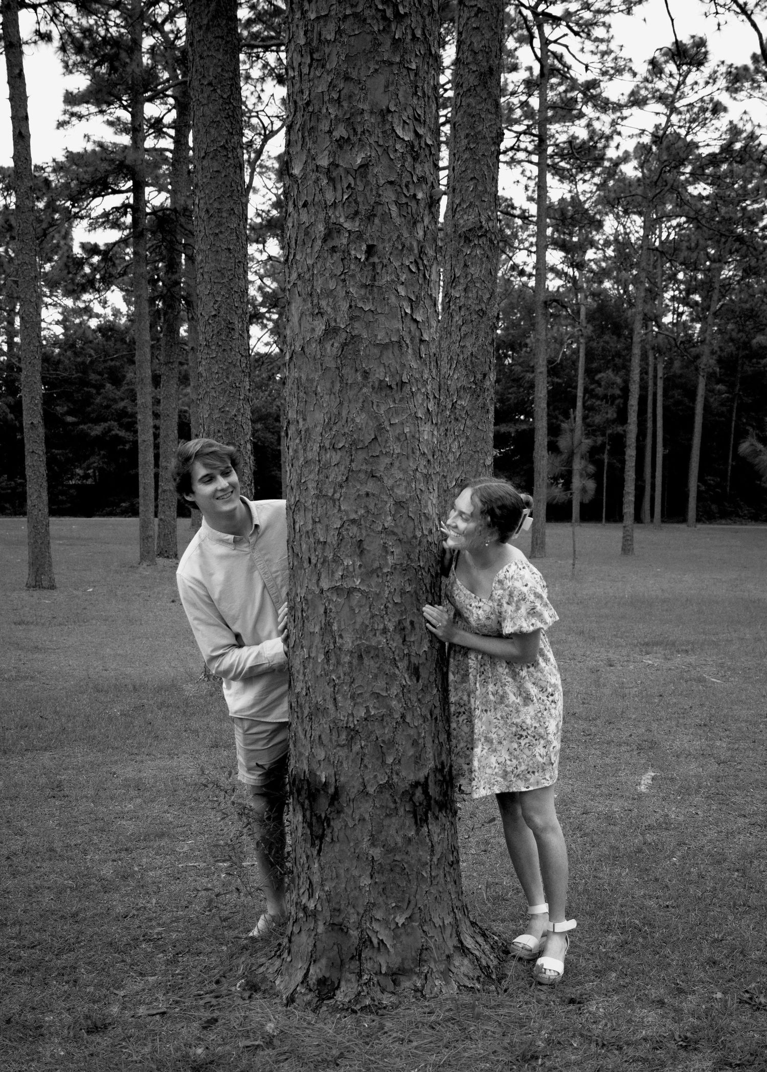 A playful black and white photo of two people on opposite sides of a large tree trunk.