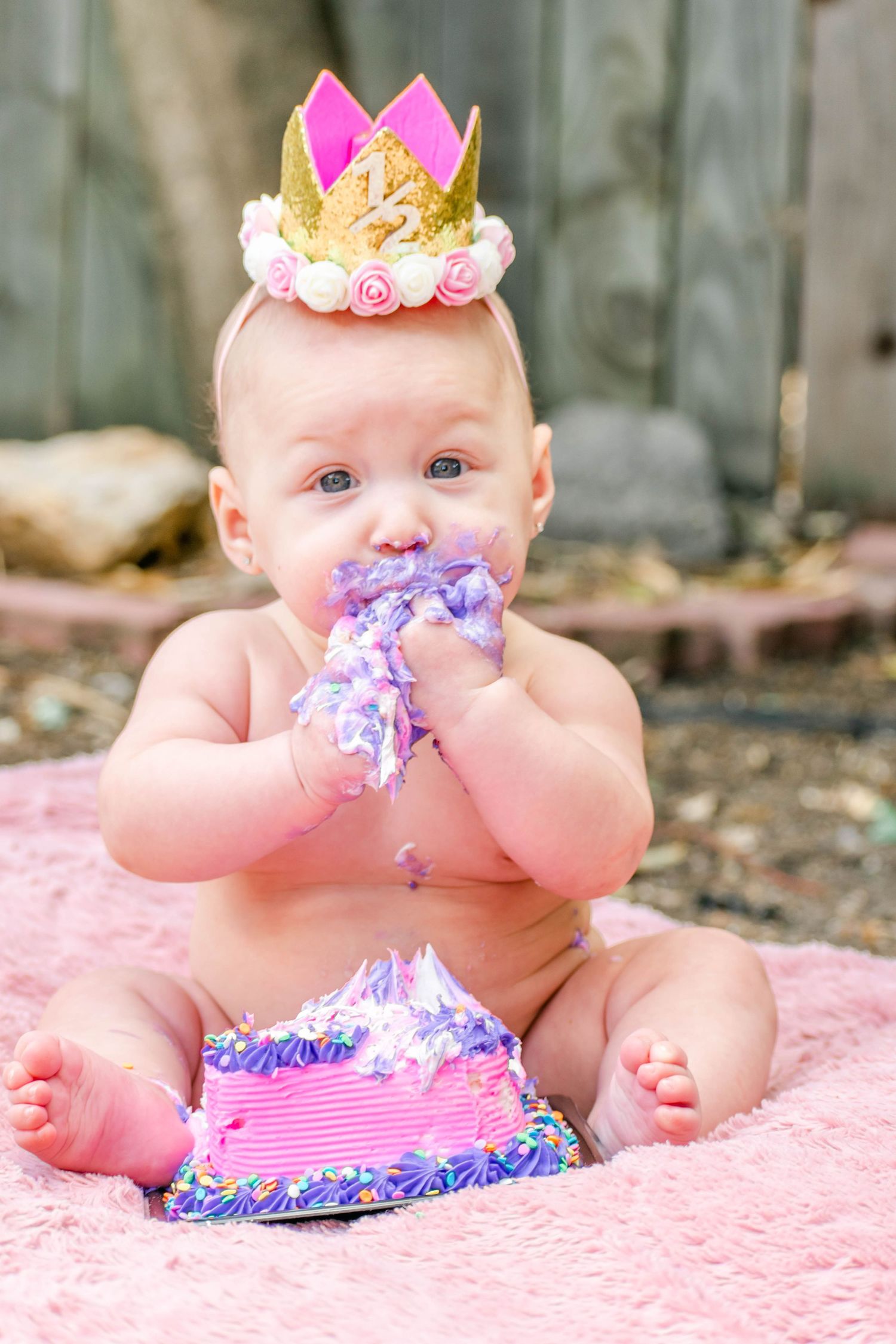 First birthday portrait of baby wearing crown playing with purple cake on pink backdrop.