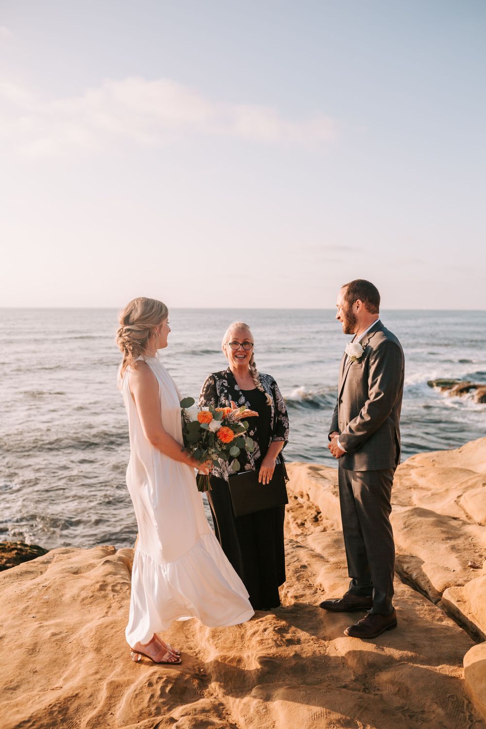A romantic coastal wedding ceremony takes place on rocky cliffs overlooking the ocean at sunset.