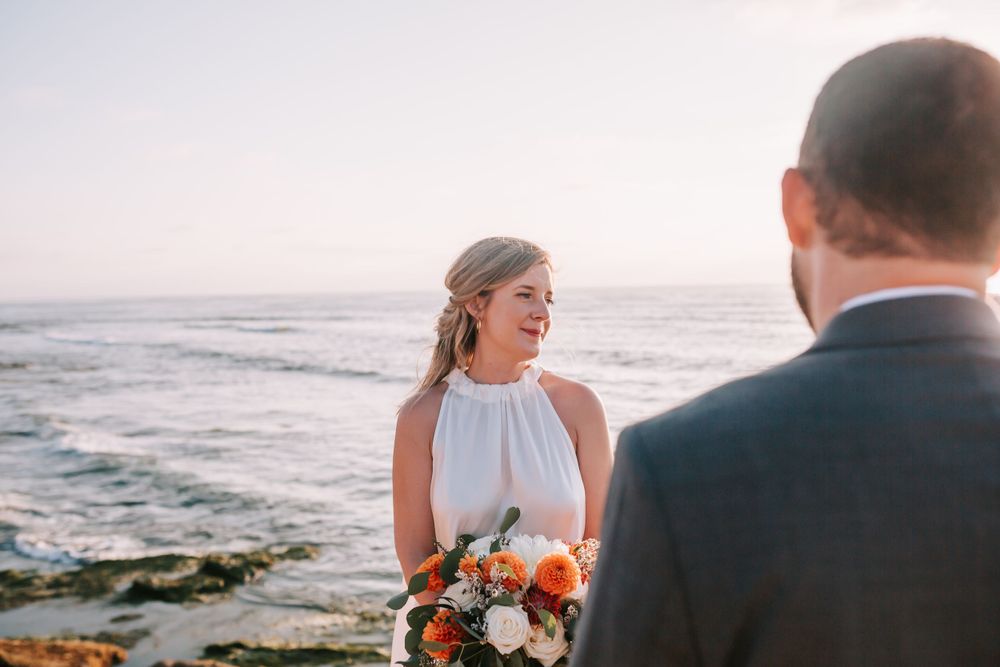A coastal wedding ceremony at sunset with a bride in a white dress holding a vibrant bouquet by the ocean.