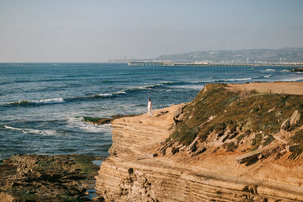 Scenic coastal cliff overlooks Pacific Ocean waves with distant pier and urban skyline visible on the horizon.