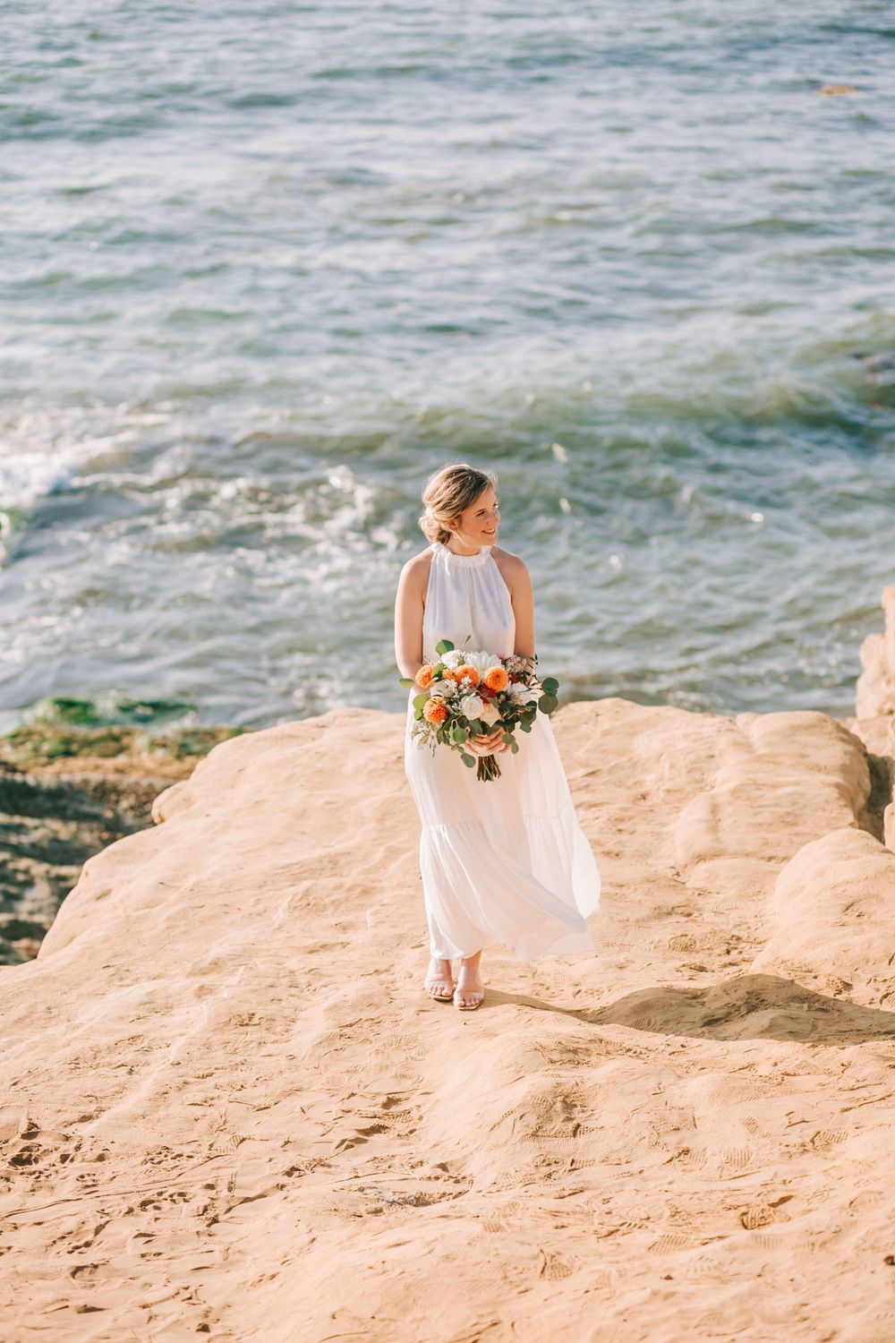 A bride in a flowing white dress holds a floral bouquet while standing on coastal rocks overlooking the ocean.