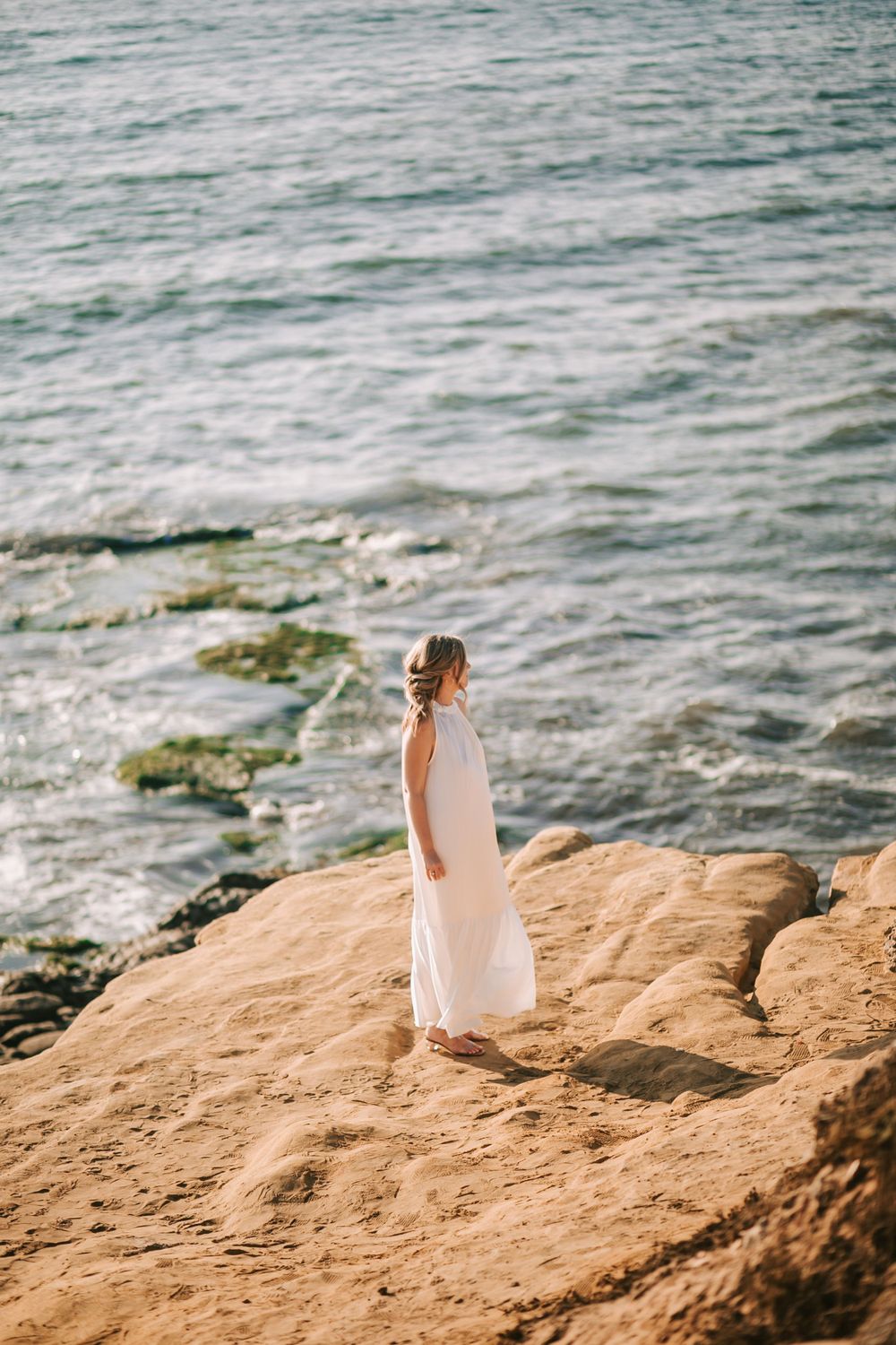 A bride stands on coastal rocks overlooking the ocean on a sunny day.