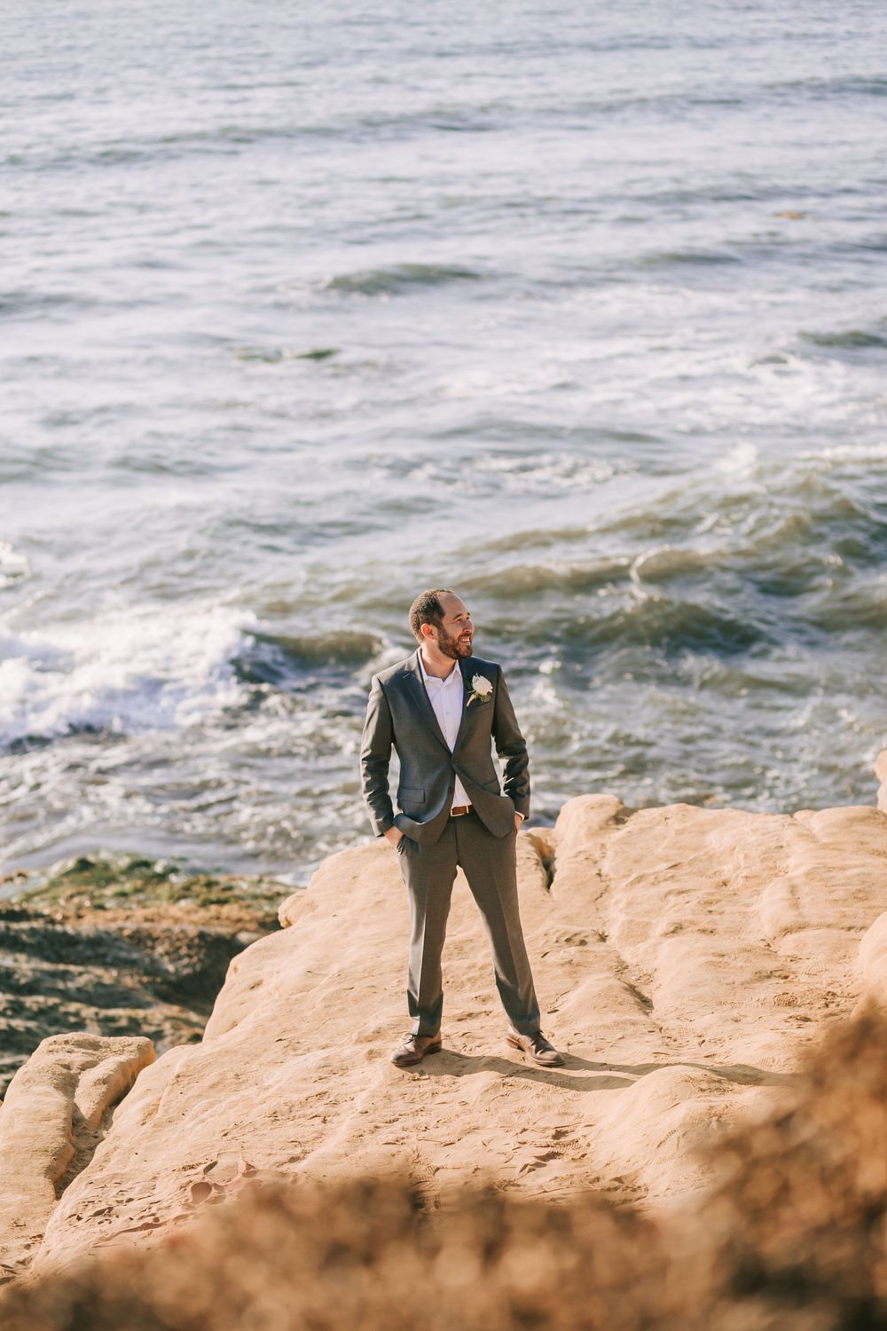 A person in a formal suit stands confidently on coastal rocks overlooking the ocean waves on a bright day.