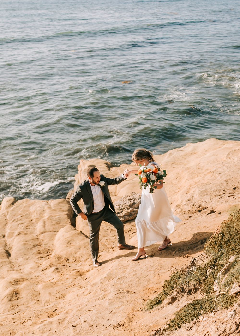 A couple in wedding attire climbs together on coastal cliffs overlooking the ocean during sunset.