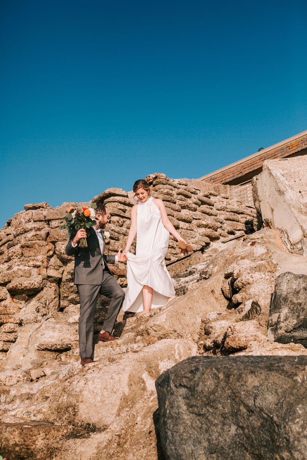 Two people in formal attire climbing rocky terrain against bright blue sky on sunny day.