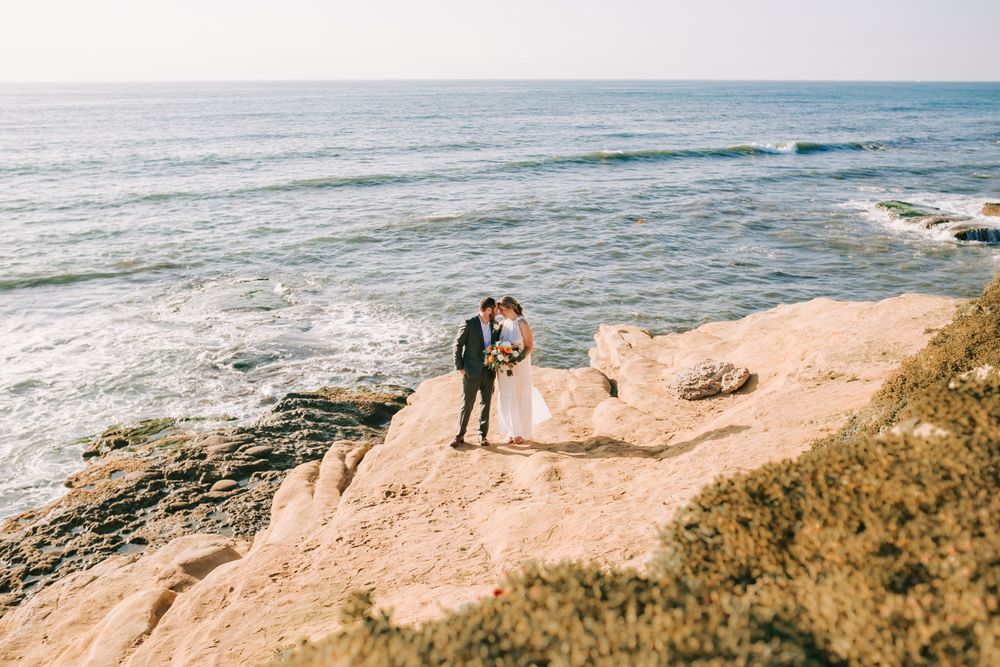 Couple stands on scenic coastal cliffs overlooking the ocean waves and rocky shoreline on a sunny day.