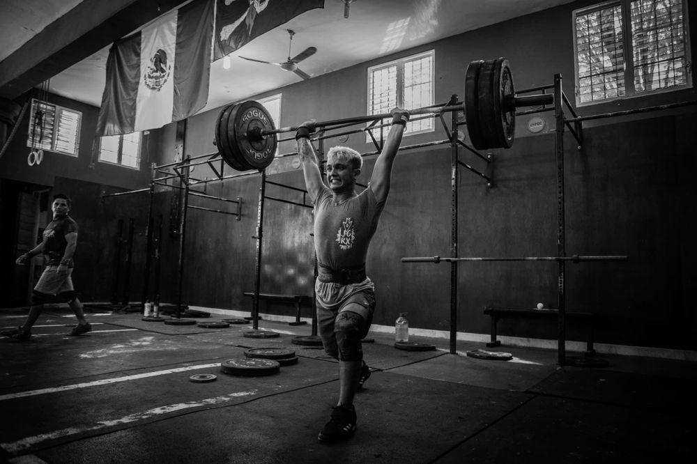 Black and white photo of an athlete performing an overhead barbell lift in a gym setting.