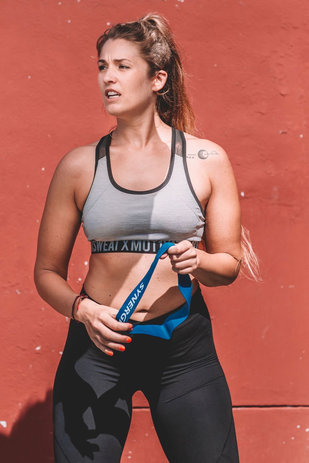 Athletic woman in workout gear holding resistance bands against an orange wall.