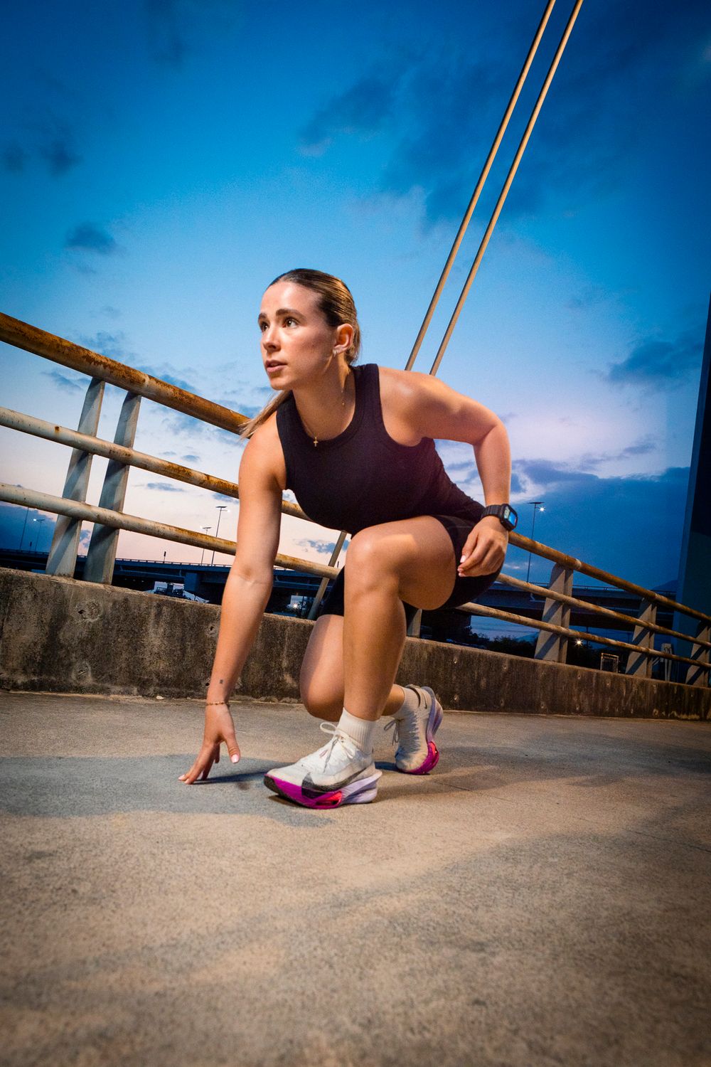 Female runner in starting position on urban bridge at dusk, wearing purple athletic wear and colorful sneakers.