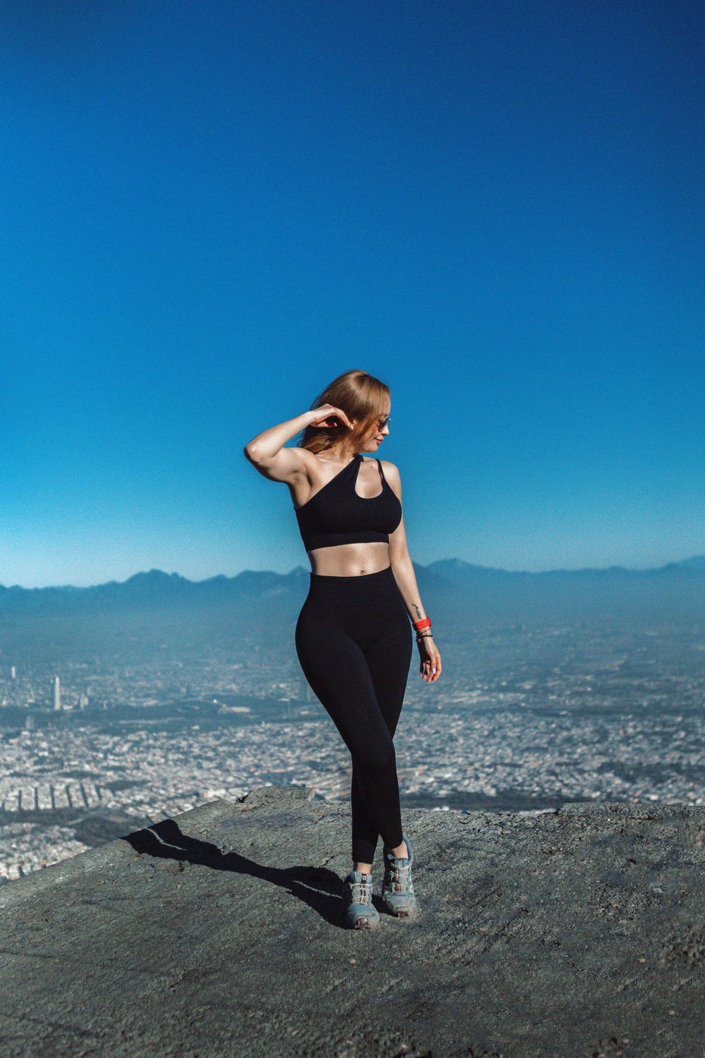 Athletic figure in black workout attire poses against cityscape backdrop on mountain overlook.