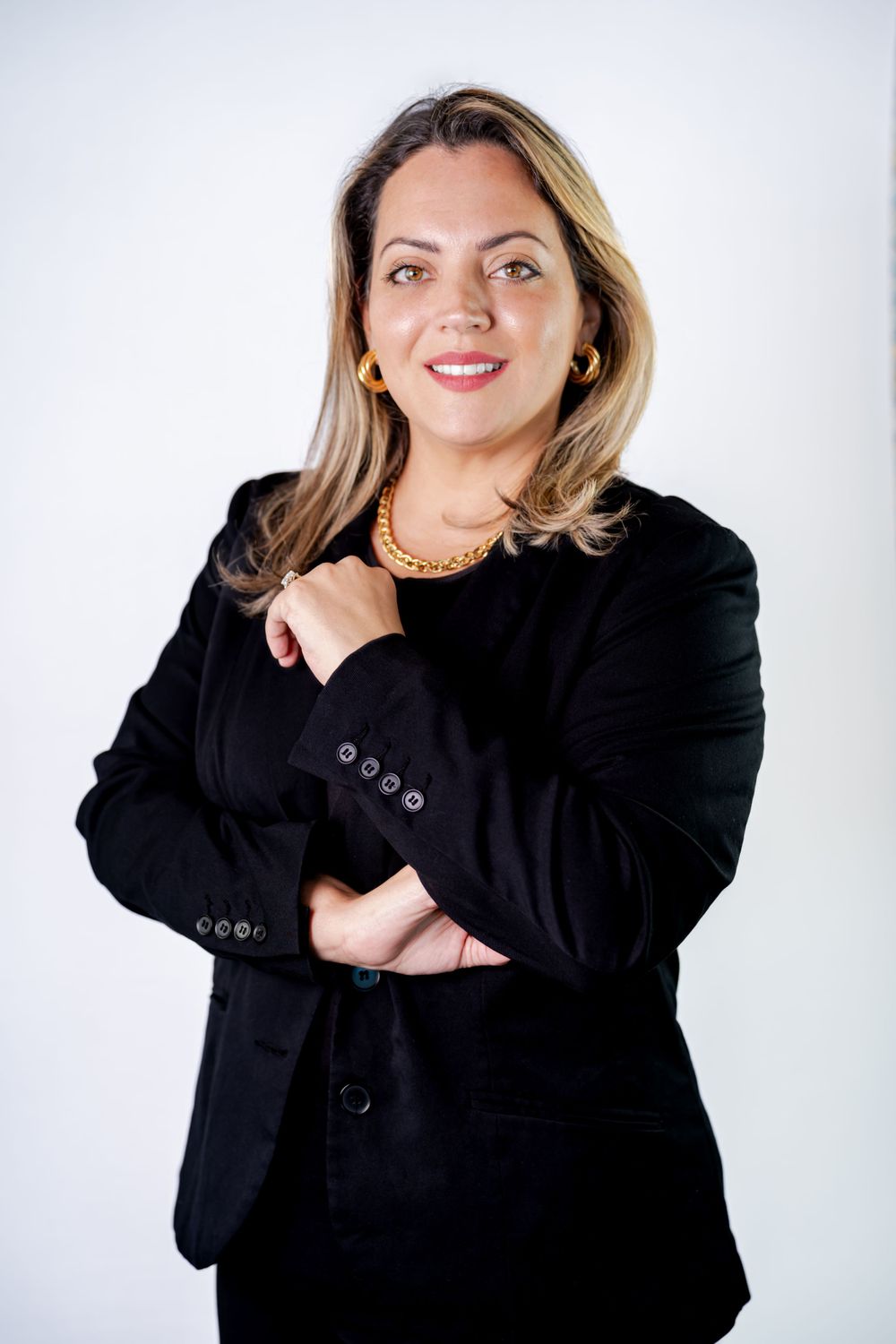 Professional portrait of someone in a black business suit and gold necklace standing against a light background.