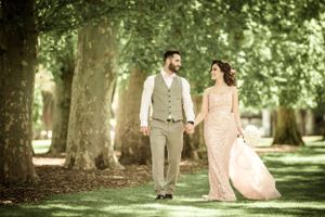 A couple walks hand in hand through a sun-dappled garden path wearing elegant attire.