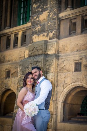 Wedding couple in formal attire posing against a historic stone building with architectural details.