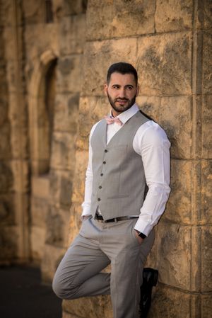 Groom in gray vest and white shirt poses against stone wall with pink bowtie.