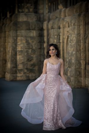Bride in blush pink sequined gown with flowing train poses against stone architecture.