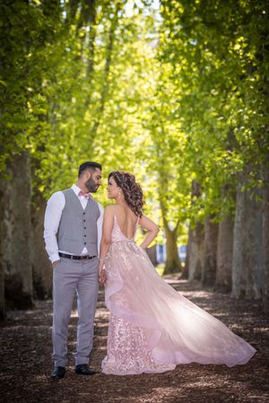A romantic wedding photo of a couple in a tree-lined pathway with the bride wearing a flowing pink lace gown.